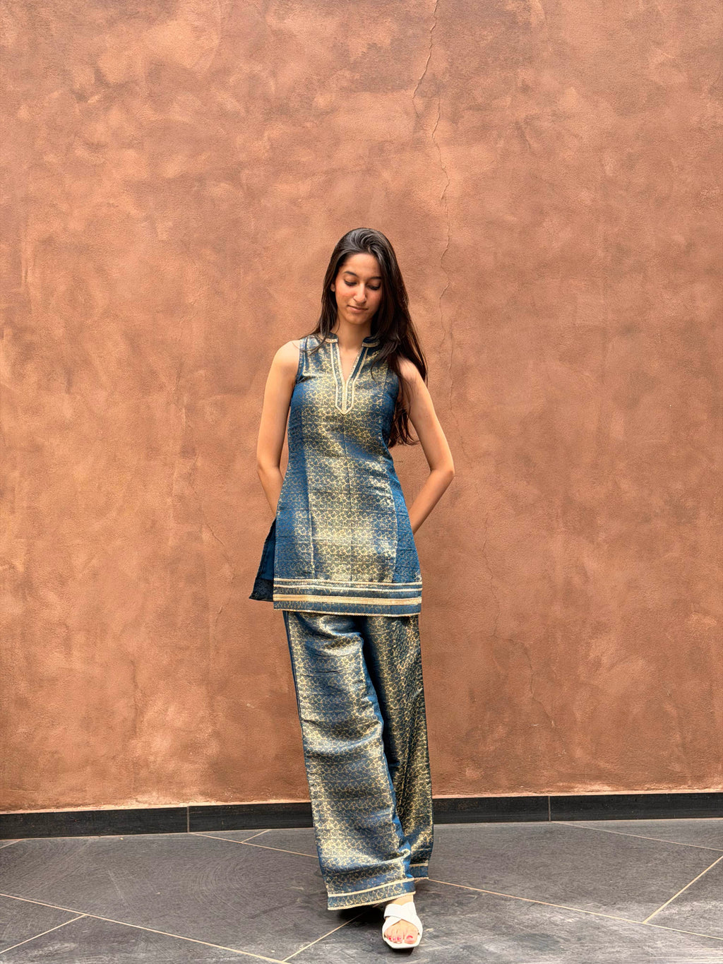A woman modeling a metallic blue and gold printed kurta set with matching wide-leg trousers, standing against a textured brown wall.