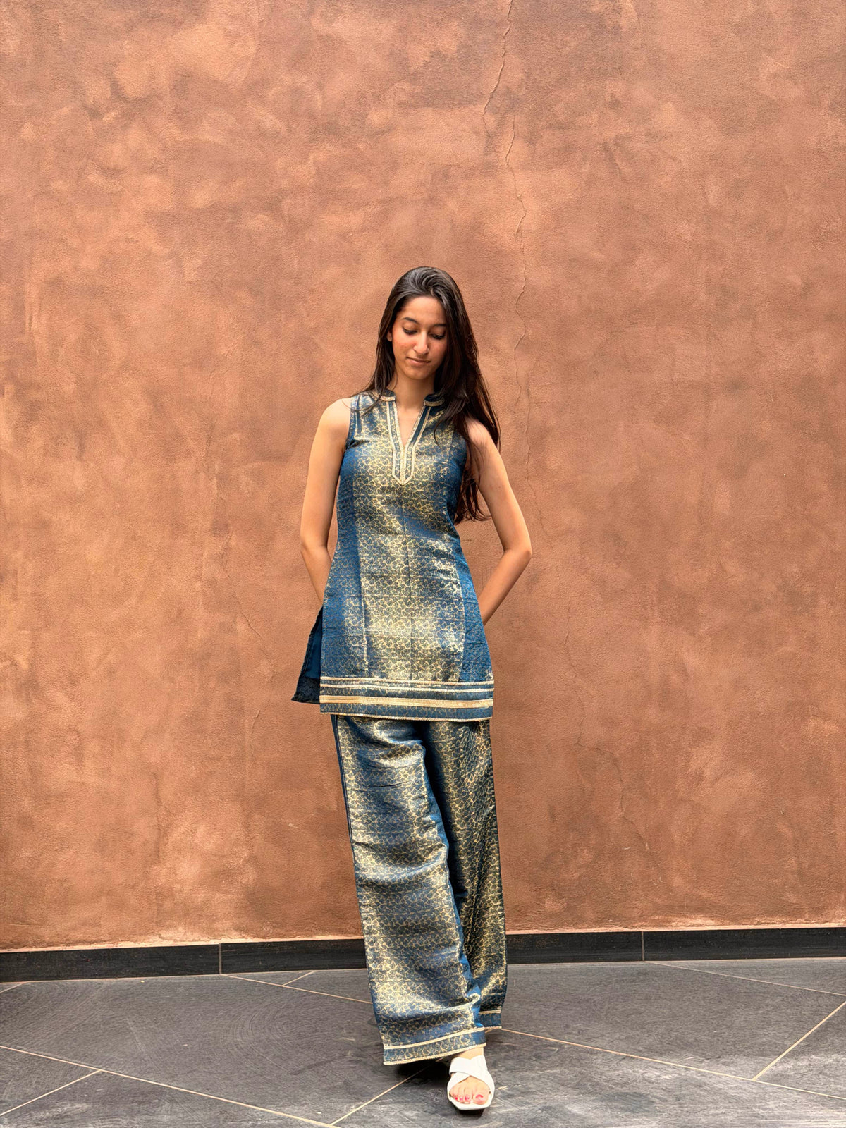 A woman modeling a metallic blue and gold printed kurta set with matching wide-leg trousers, standing against a textured brown wall.
