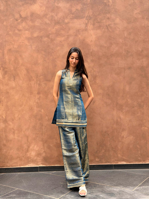 A woman modeling a metallic blue and gold printed kurta set with matching wide-leg trousers, standing against a textured brown wall.