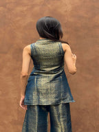 A woman viewed from behind wearing a sleeveless metallic patterned tunic and matching wide-leg pants against a textured brown wall.