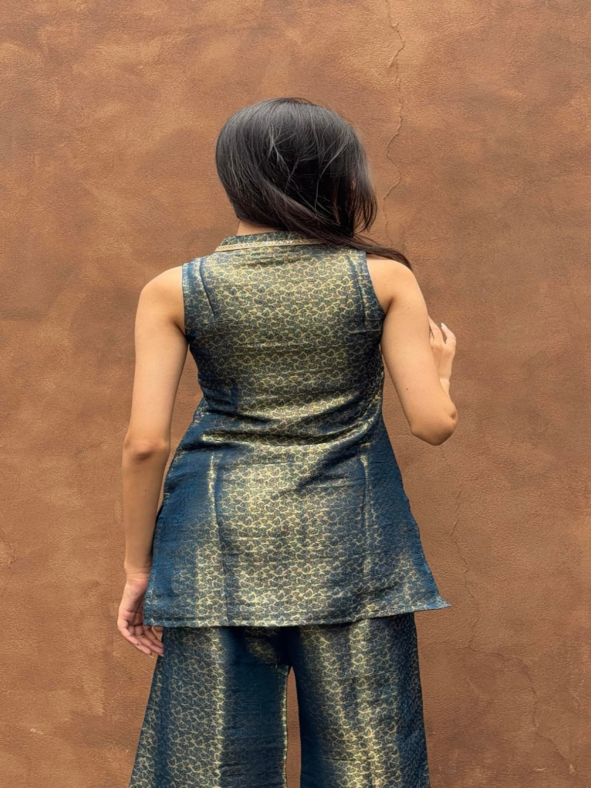 A woman viewed from behind wearing a sleeveless metallic patterned tunic and matching wide-leg pants against a textured brown wall.