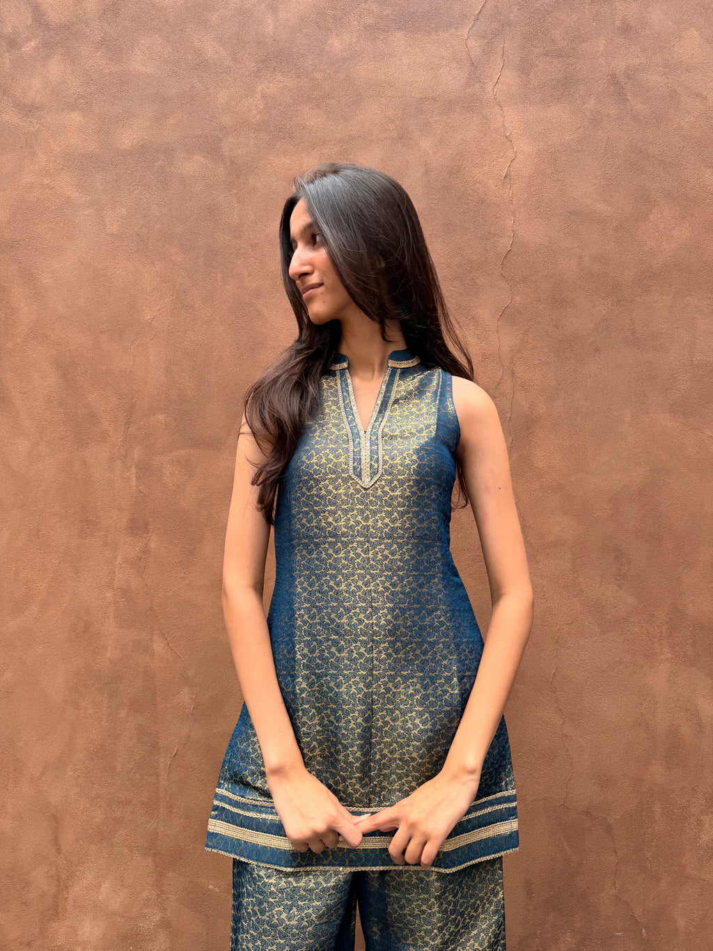 A woman modeling a sleeveless blue and gold patterned kurta with matching pants, standing against a textured brown stucco wall.