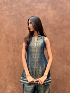A woman modeling a sleeveless blue and gold patterned kurta with matching pants, standing against a textured brown stucco wall.