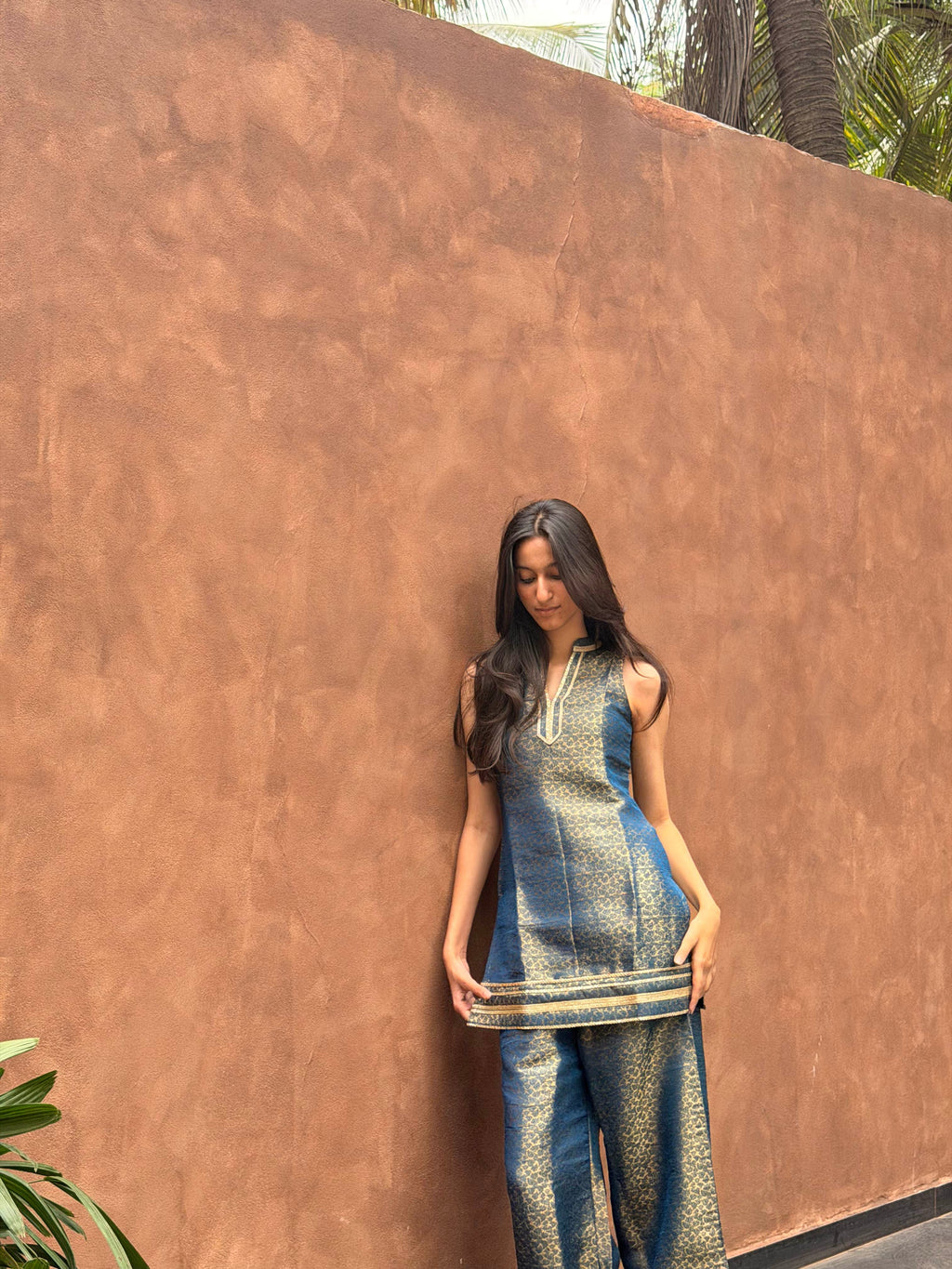 A woman stands against a large textured terracotta wall wearing a blue and gold patterned kurta set with matching wide-leg pants.
