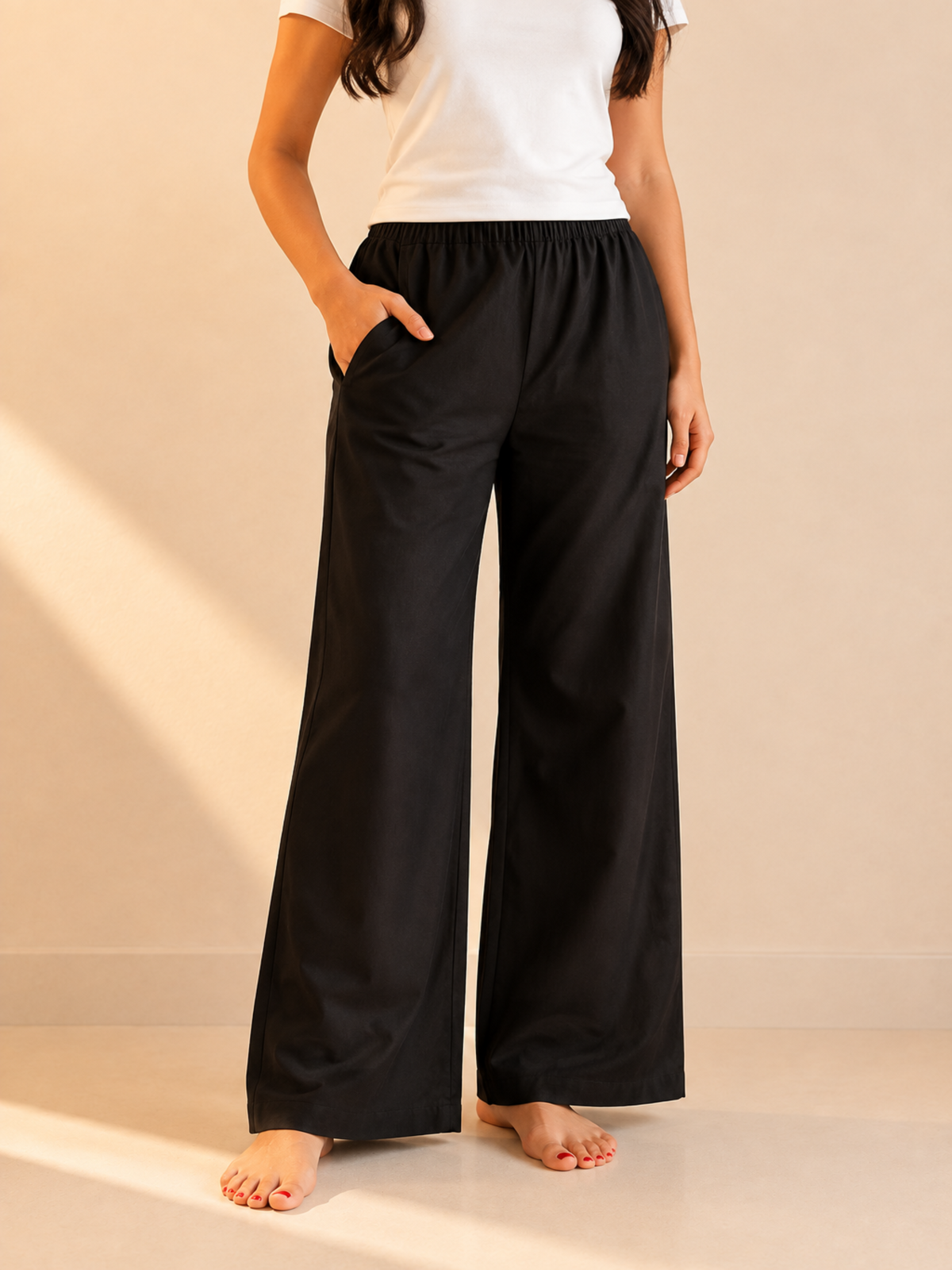 A person wearing black wide-leg lounge pants with a white top, standing barefoot against a warm beige wall with sunlight casting soft shadows.