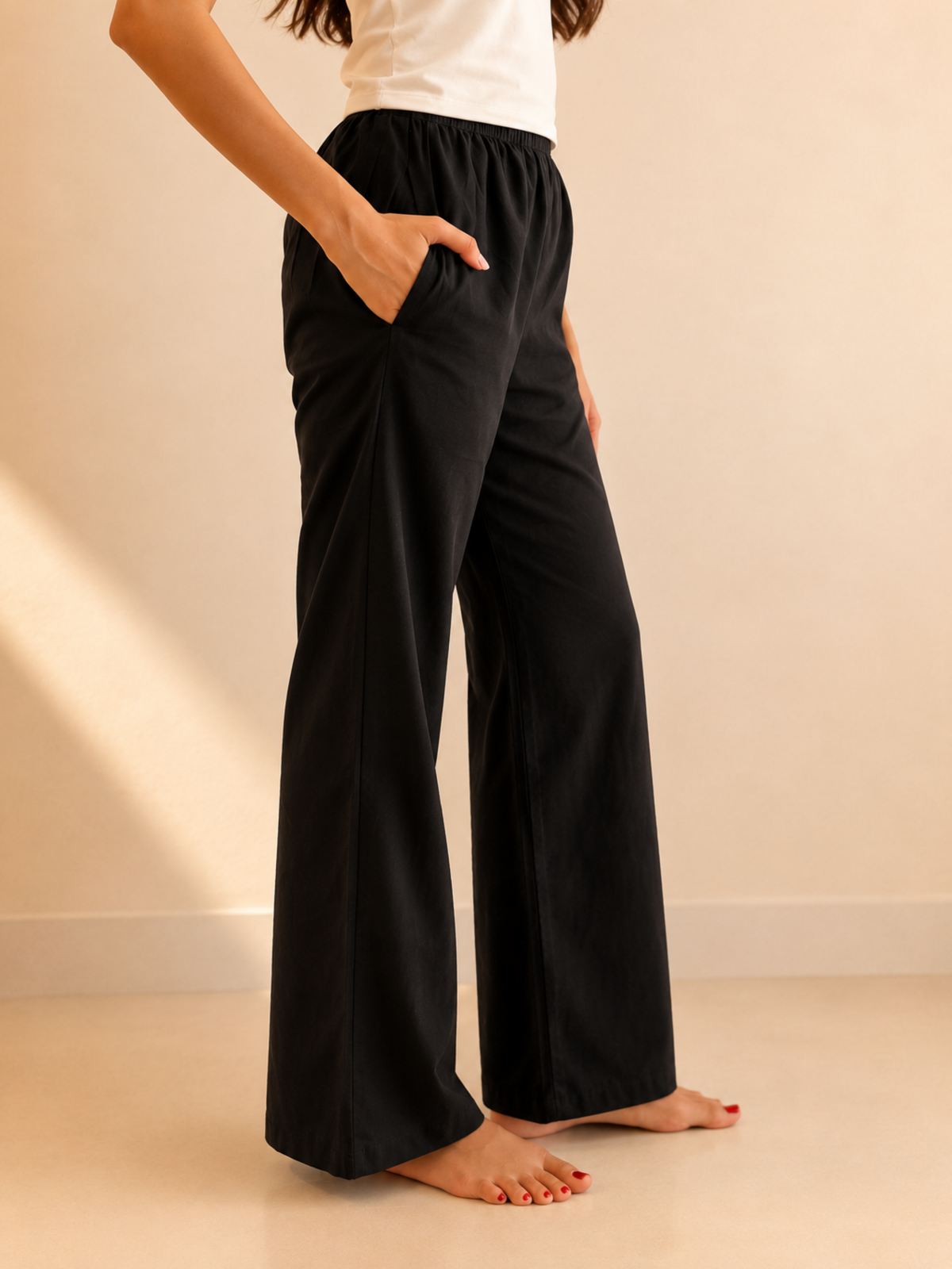 A person wearing black wide-leg lounge pants with side pockets, paired with a white top, standing barefoot against a warm beige wall and floor.
