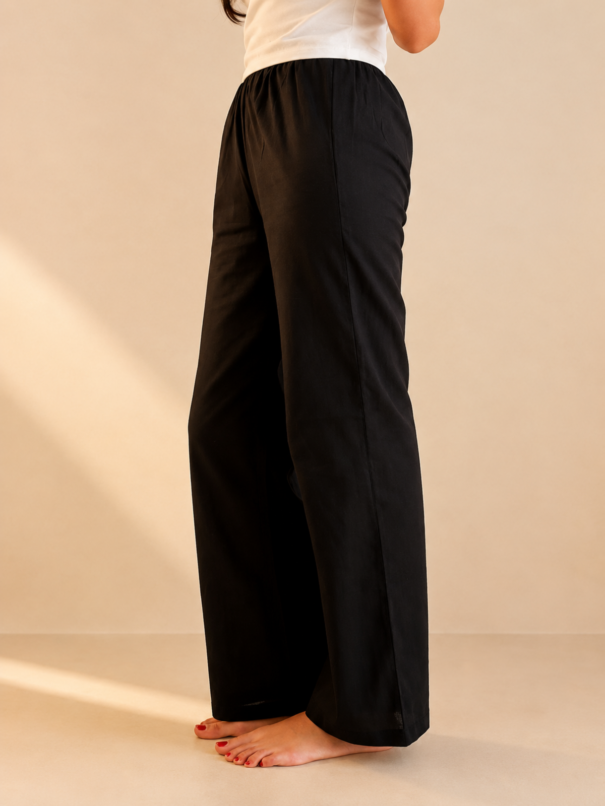 A person wearing loose black wide-leg pants and a white top, standing barefoot against a warm beige studio background.