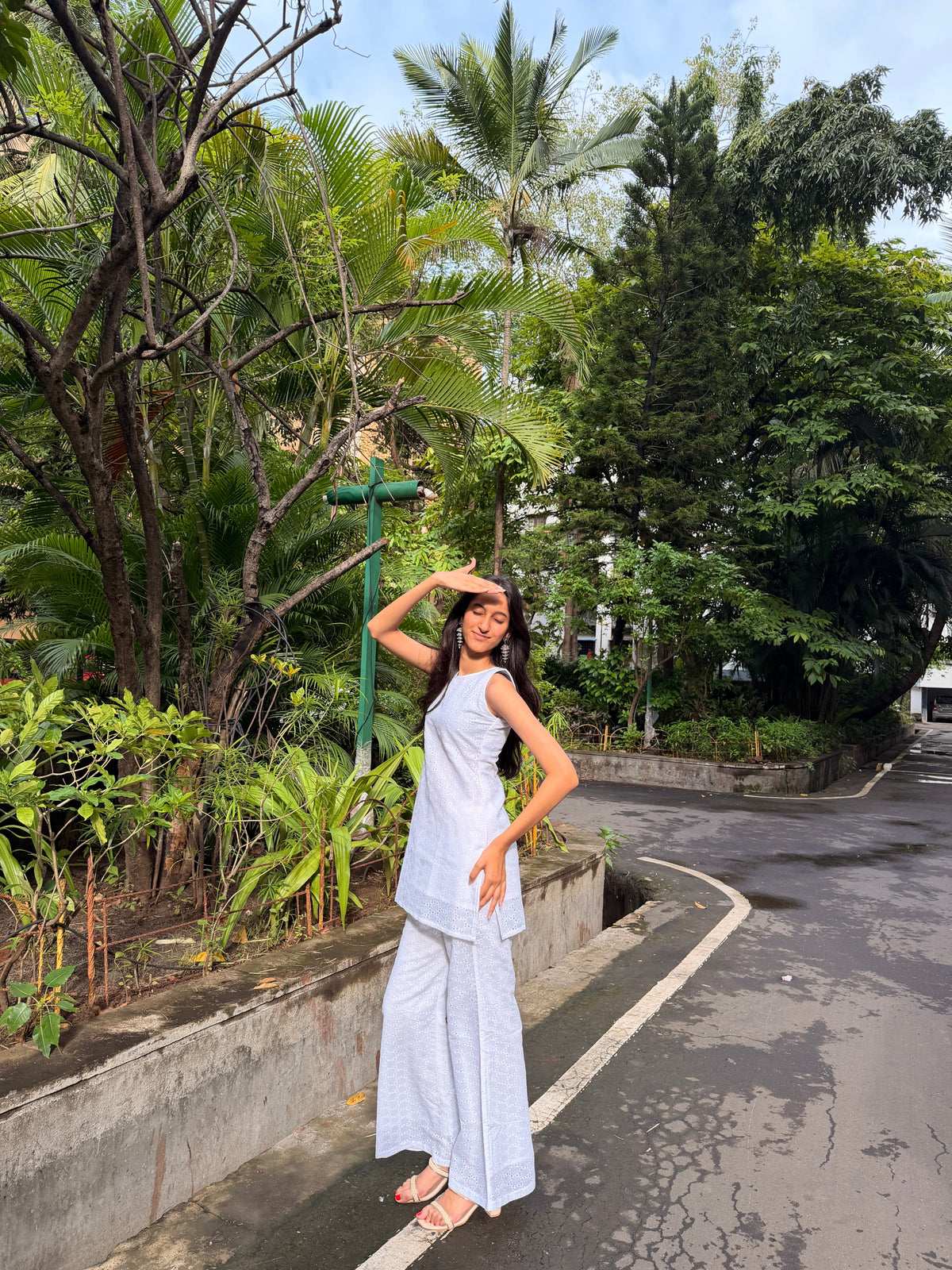 A woman posing outdoors on a paved road lined with lush tropical greenery, wearing a light blue and white patterned sleeveless tunic top and matching wide-leg pants.