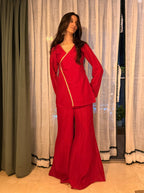 A woman modeling a red two-piece matching set consisting of a long-sleeve wrap-style top with a gold zipper detail and matching wide-leg pants, standing in front of white and green curtains.
