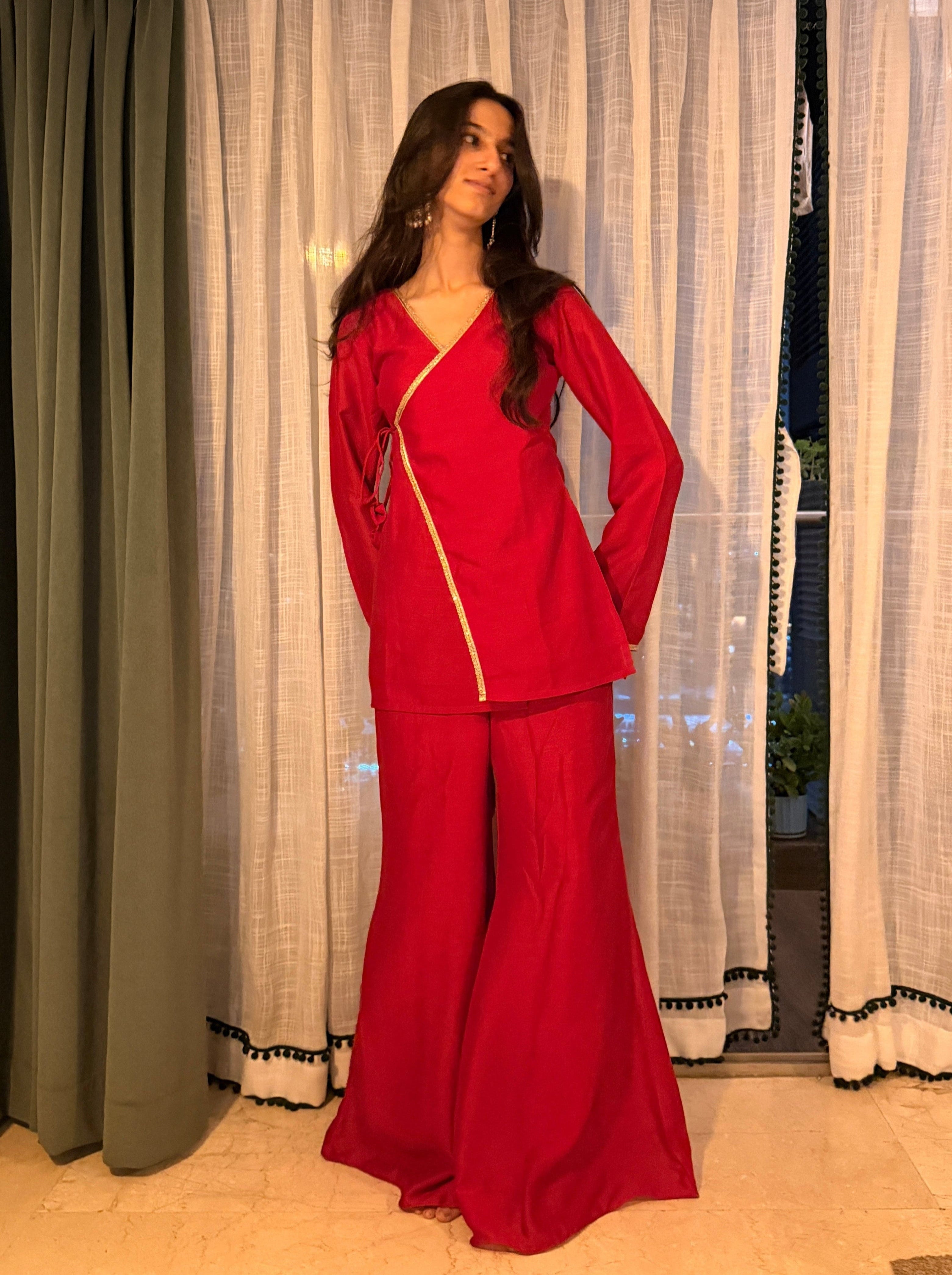 A woman modeling a red two-piece matching set consisting of a long-sleeve wrap-style top with a gold zipper detail and matching wide-leg pants, standing in front of white and green curtains.