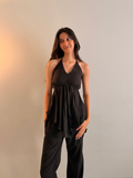 A woman modeling a black sleeveless halter top with a gathered peplum-style ruffle at the waist, paired with black wide-leg pants, standing against a plain light gray wall.