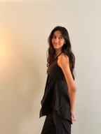 A woman with long dark hair is wearing a black sleeveless top and black pants, posing against a plain off-white wall with a warm light on the left.