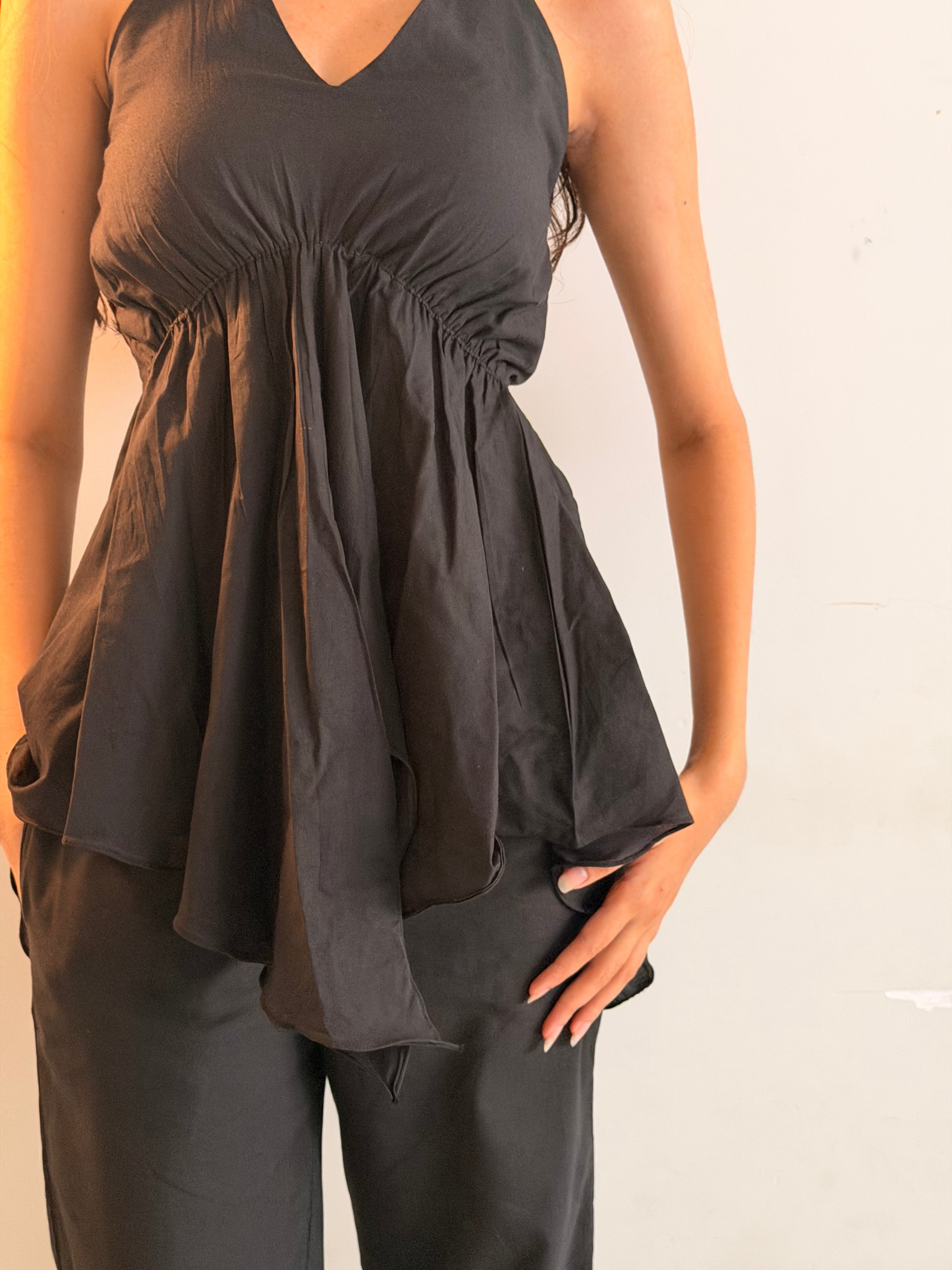A woman wearing a black sleeveless peplum-style top with a gathered waist and matching black pants, standing against a plain light wall.