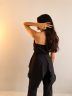 A woman with long dark hair is shown from behind wearing a black strapless top and matching black pants against a warm beige studio background.