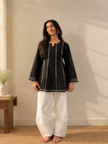 A woman stands barefoot indoors wearing a black long-sleeve kurta with white contrast stitching and white wide-leg pants with gold detailing near the hem. The room has a beige wall, a small wooden side table with a vase, and natural light casting shadows on the wall.