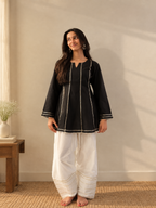 A woman stands barefoot indoors wearing a black long-sleeve kurta with white contrast stitching and white wide-leg pants with gold detailing near the hem. The room has a beige wall, a small wooden side table with a vase, and natural light casting shadows on the wall.