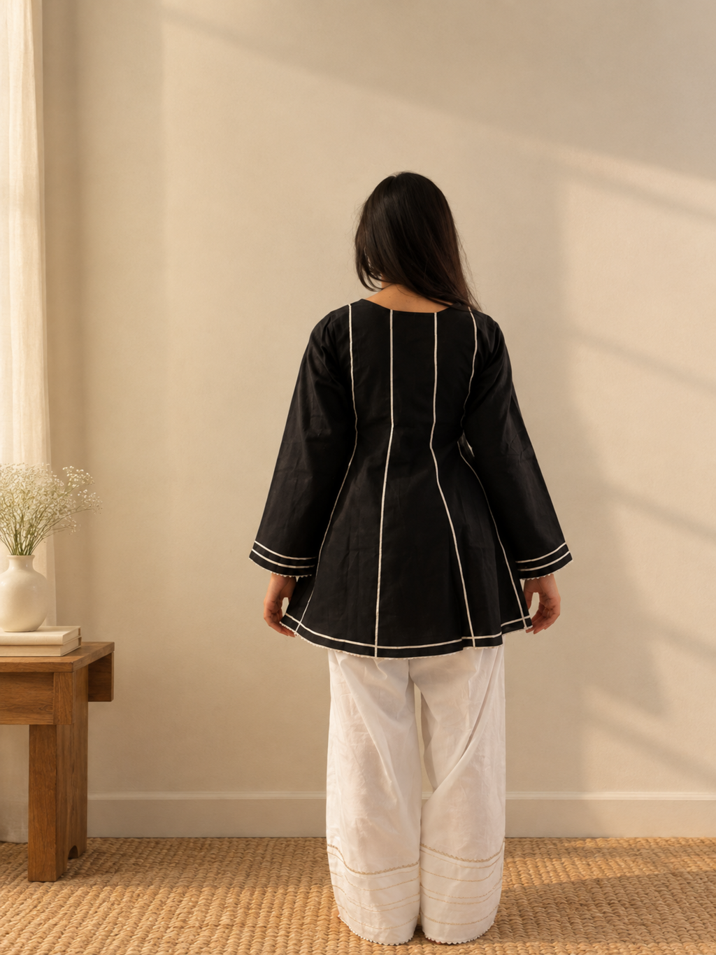 A woman stands with her back to the camera wearing a black long-sleeve tunic top with white contrast stitching and white wide-leg pants in a sunlit room.