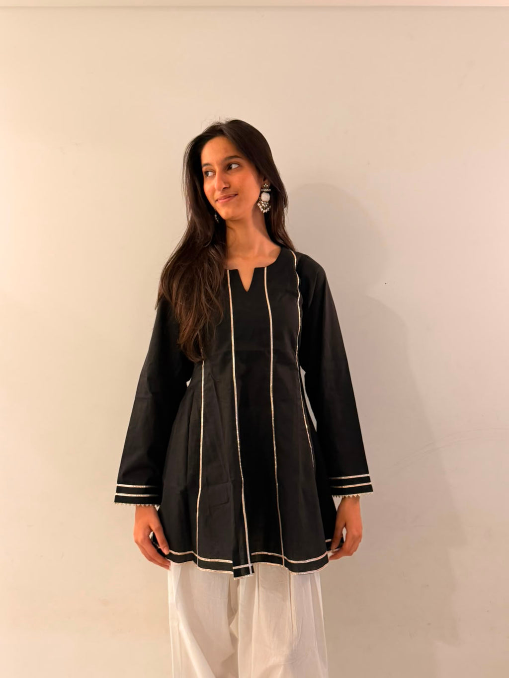 A woman modeling a black long-sleeve kurta with white contrast stitching and piping details, paired with white pants, standing against a plain off-white wall.