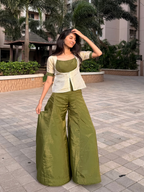 A woman modeling an olive green traditional outfit consisting of wide-leg trousers and a matching top with a cream-colored embroidered jacket, standing outdoors on a paved walkway.