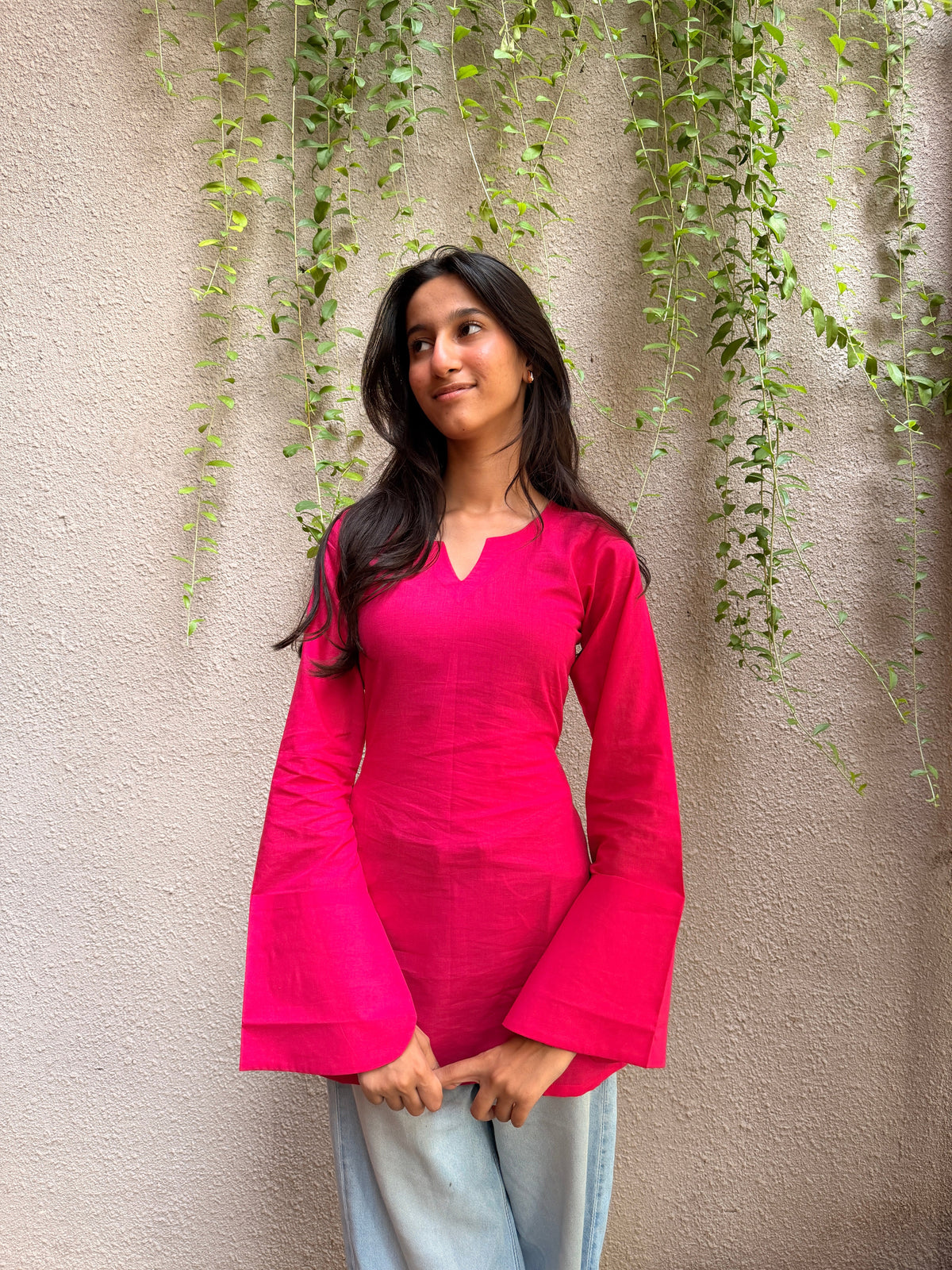 A woman wearing a bright pink long-sleeve top with bell sleeves and light blue jeans, standing against a textured beige wall with green trailing vines.