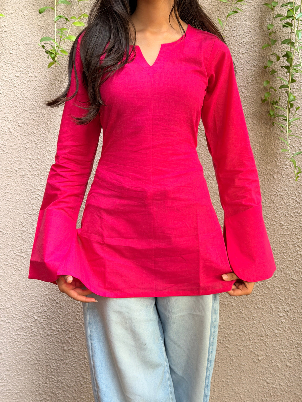 A woman wearing a bright fuchsia pink long-sleeve tunic top with a split V-neckline and flared cuffs, paired with light blue jeans, standing against a textured beige wall with green vines.