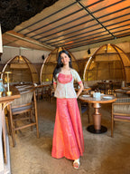 A woman stands in a restaurant wearing a traditional Thai outfit consisting of a light gray sleeveless top with pink trim and wide, flowing coral-orange skirt. The restaurant features warm wood and bamboo decor, arched seating pods, and tables with plates.