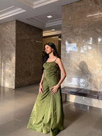 A woman wearing a floor-length olive green satin gown stands in a modern indoor lobby with marble walls and a bench.