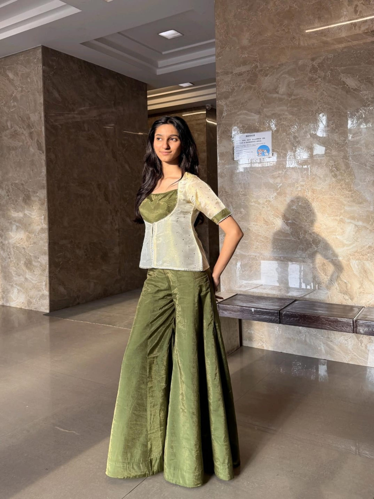 A woman models a two-piece outfit consisting of an olive green and white embroidered top paired with matching olive green wide-leg trousers, standing in a modern lobby with marble walls.